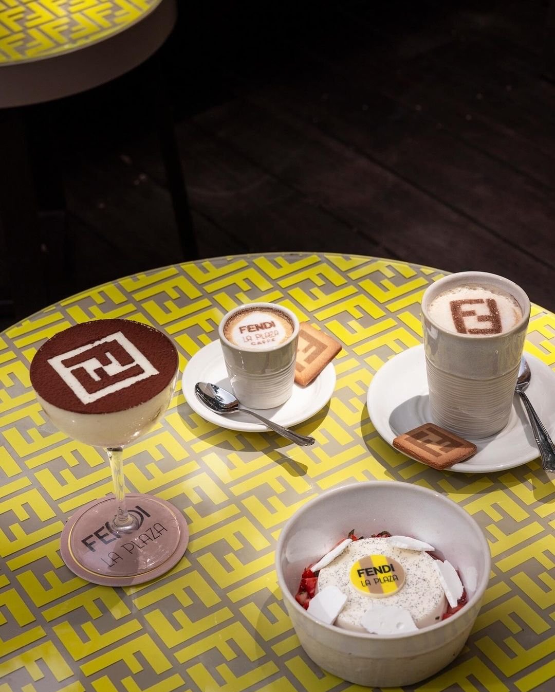 Mesa com itens de confeitaria personalizados com logotipos da grife Fendi. Cappuccinos, sobremesa e biscoito estilizados com o monograma da marca, sobre uma mesa com padrão geométrico nas cores amarelo e cinza, destacam a tendência de luxo e exclusividade.