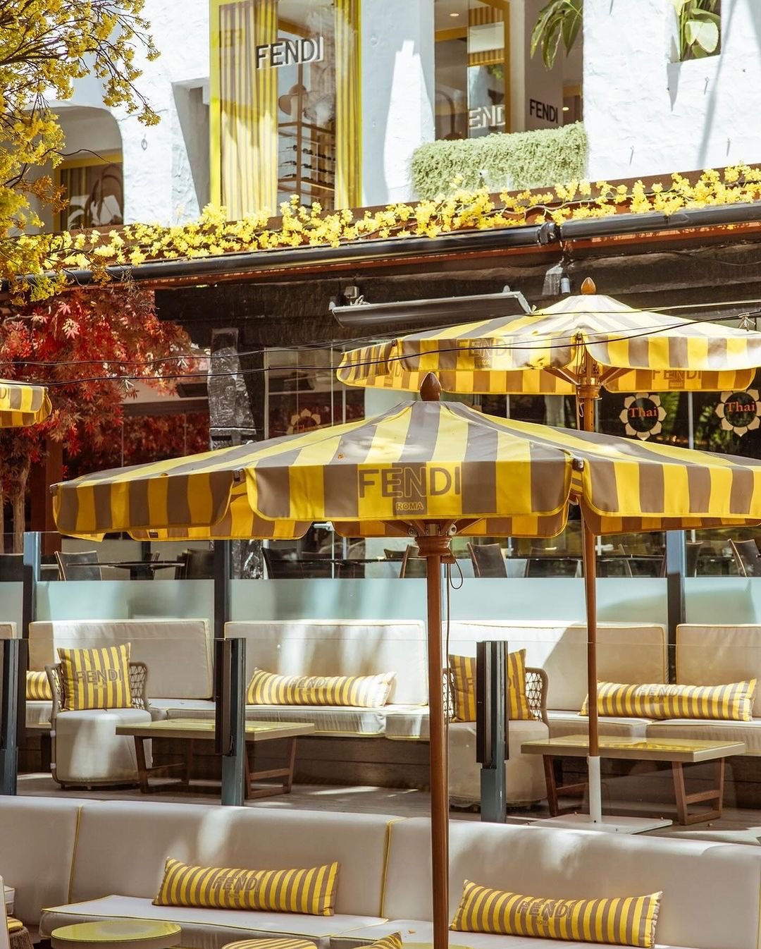 Terraço ao ar livre decorado com temática Fendi. Guarda-sóis listrados amarelo e cinza, almofadas combinando e ambiente acolhedor. Tendência de moda: luxo e sofisticação na decoração de exteriores com inspiração em grifes de alta costura.