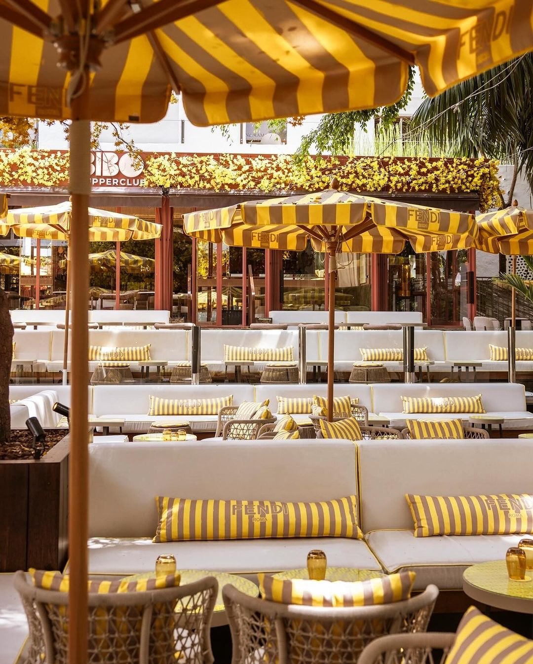 Imagem de um restaurante com decoração elegante, destacando almofadas e guarda-sóis listrados em amarelo e branco, acentuados com o logotipo da Fendi. O ambiente apresenta design sofisticado, com móveis de luxo que seguem tendências de moda e estilo contemporâneo.