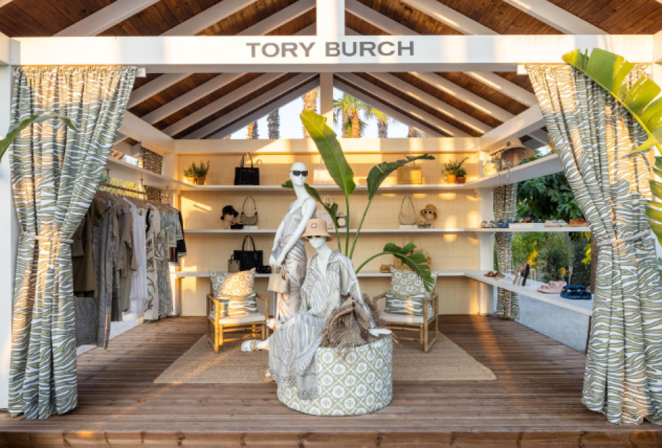 Exposição de moda Tory Burch em ambiente rústico e tropical, destacando roupas leves e acessórios sofisticados, como chapéus e bolsas elegantes. A paleta de cores neutras e estampas naturais enfatizam o estilo elegante e descontraído, ideal para climas quentes. verão europeu 