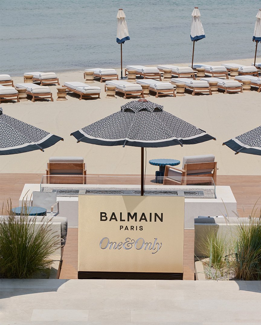 Praia sofisticada com espreguiçadeiras de madeira e colchões brancos, guarda-sóis estampados com logomarca Balmain. Tendência de moda praia de luxo, destacando estilo e design exclusivo. Combinação de elegância e conforto em ambiente à beira-mar. verão europeu