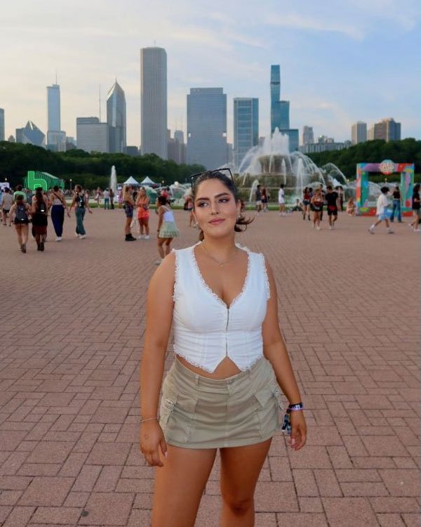 Mulher com top branco de renda e mini-saia bege, posando em um local urbano com arranha-céus ao fundo. A roupa destaca tendências de moda casual e verão, com um toque feminino e elegante.