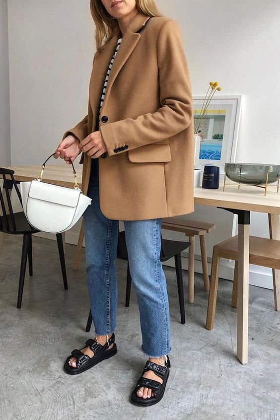 Look casual-chique com blazer caramelizado oversized, calça jeans reta, camiseta listrada e sandálias pretas confortáveis. A bolsa branca de mão complementa a combinação. Perfeito para um visual moderno e descontraído com toques clássicos.