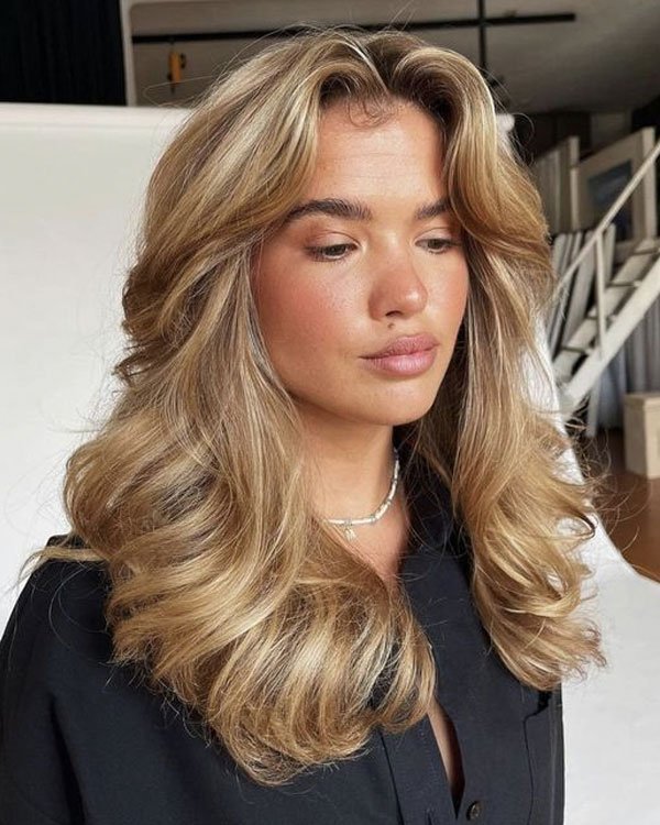 Imagem de mulher com cabelo loiro volumoso e ondas suaves, vestindo camisa preta de tecido leve e colar discreto.