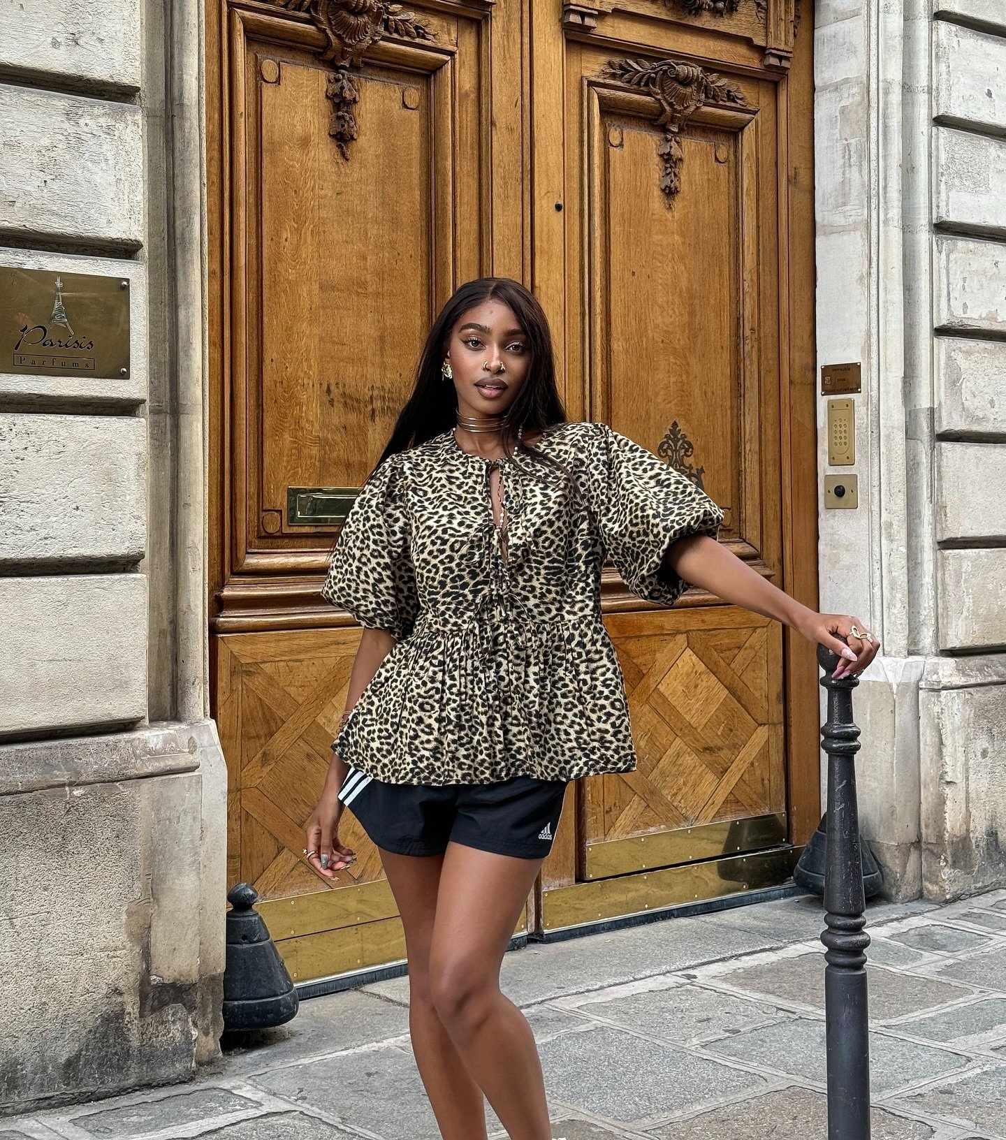 Mulher vestindo uma blusa com estampa de leopardo e mangas bufantes, combinada com shorts esportivos pretos da marca Adidas. Look casual misturando elementos de moda clássica animal print com tendências esportivas, ideal para um estilo urbano despojado.