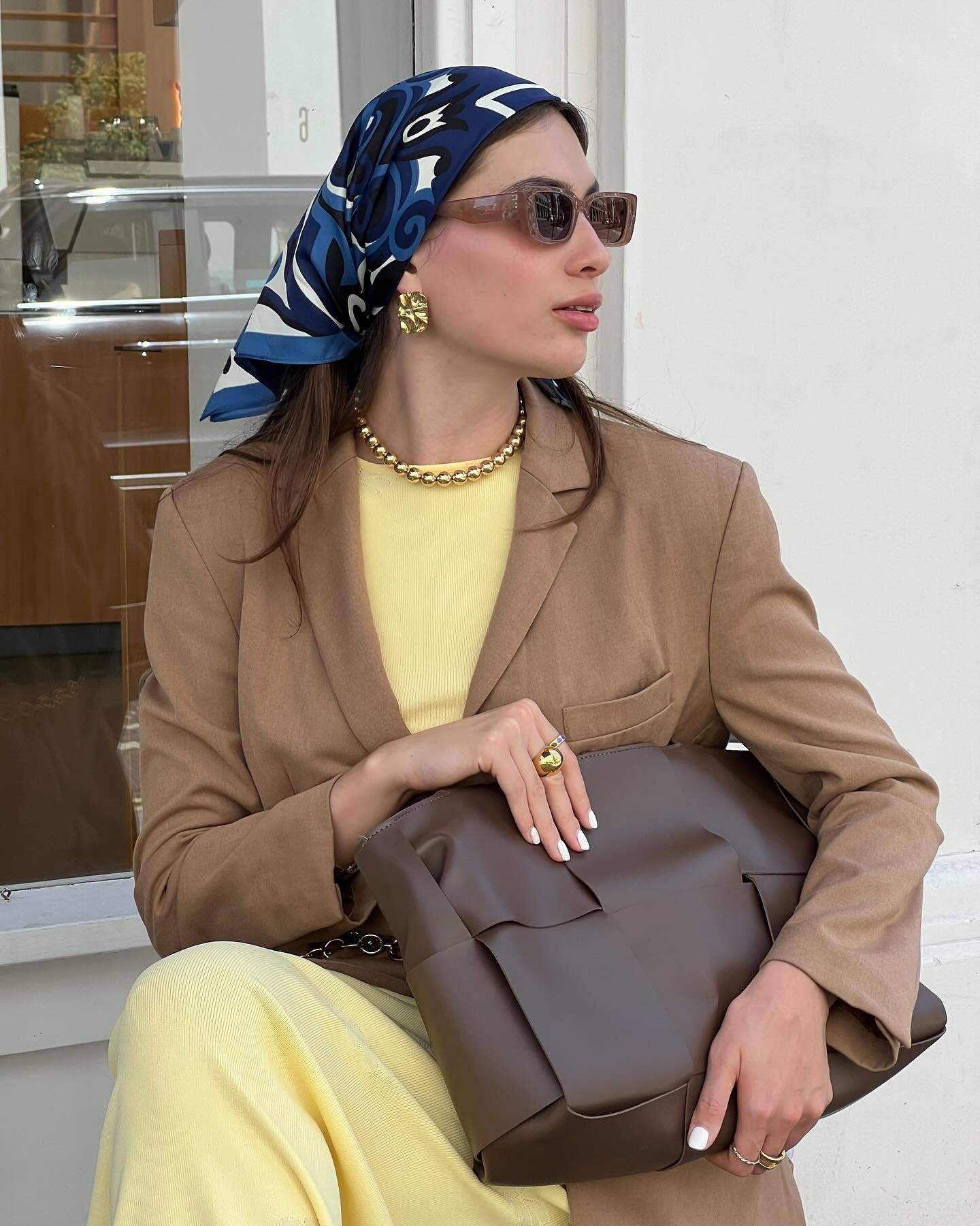 Mulher elegante usando lenço azul estampado na cabeça, óculos de sol quadrados, blazer bege, vestido amarelo claro, colar de pérolas, anéis dourados com detalhes, carregando bolsa marrom grande. Look clássico e sofisticado, ideal para moda urbana e contemporânea.