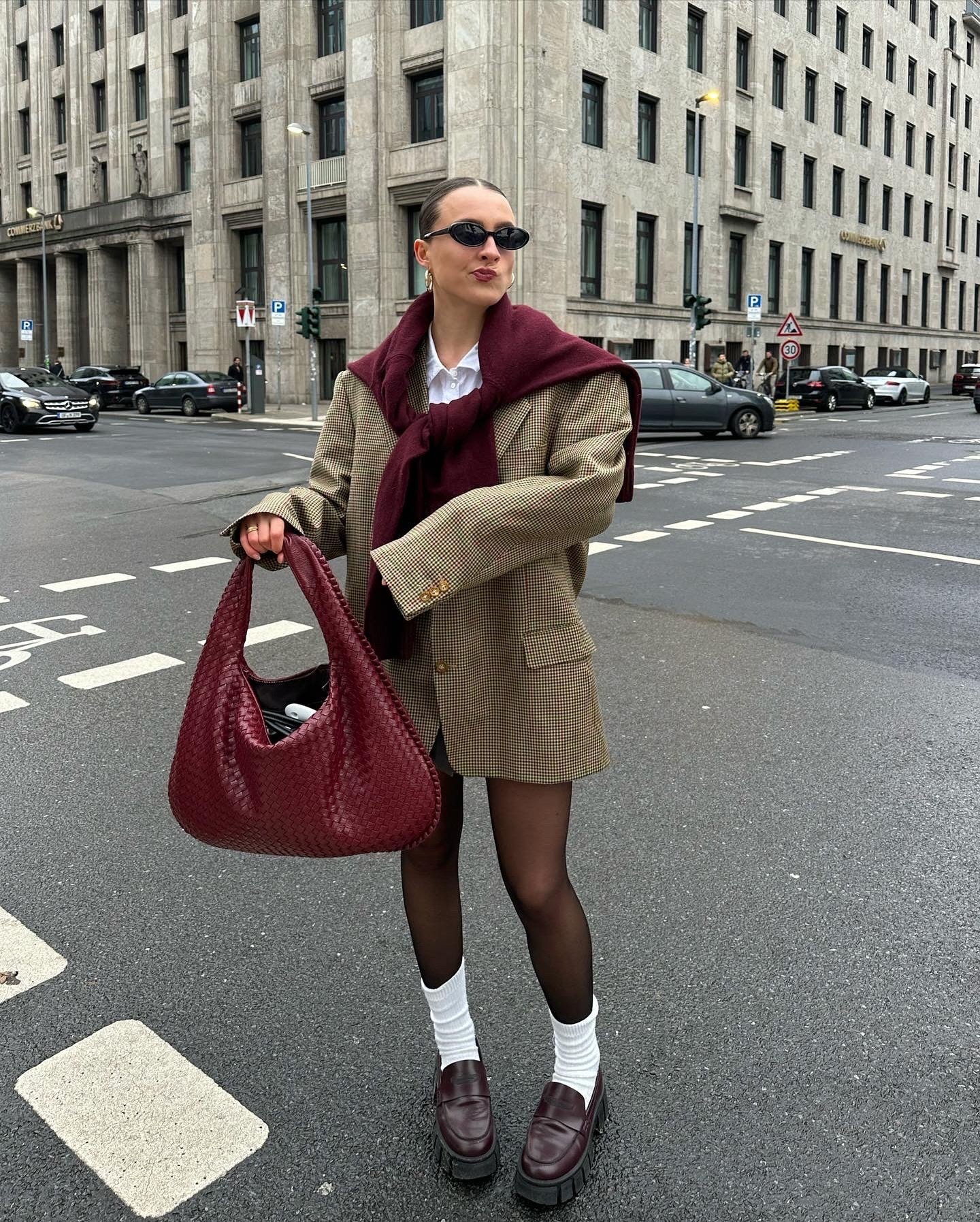 Mulher vestida com blazer oversized xadrez, suéter vinho nos ombros, meias brancas, meia-calça preta e loafers vinho de sola tratorada. Carrega bolsa de couro vinho trançado e usa óculos de sol pequenos. Estilo elegante e moderno, ideal para o outono.