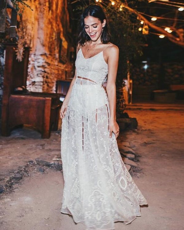 Mulher usando um vestido branco longo, transparente e rendado, com detalhes florais e recortes laterais. O estilo boho chic combina elegância e descontração, ideal para eventos noturnos ou festas ao ar livre.