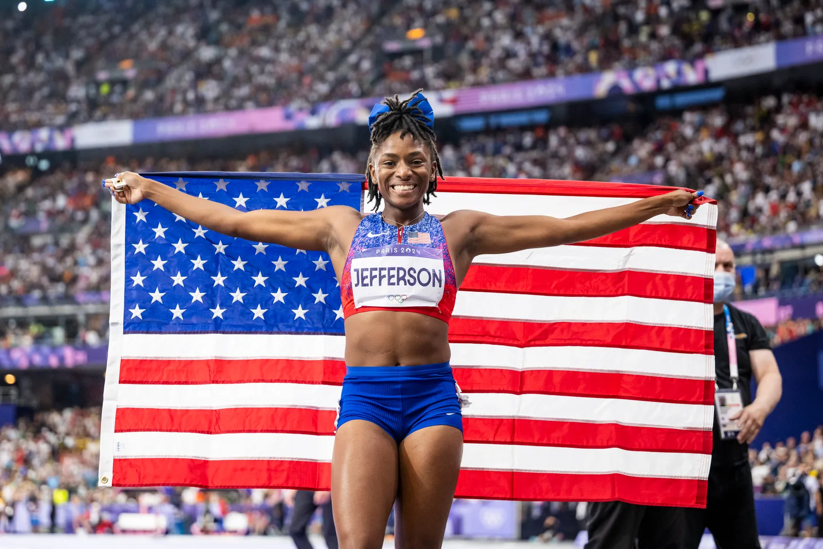 Atleta vestida em traje esportivo azul e vermelho, com top cropped e shorts curtos, exibindo a bandeira dos EUA. O look destaca conforto e funcionalidade, ideal para competições. Tendência de moda esportiva,
olimpíadas 2024