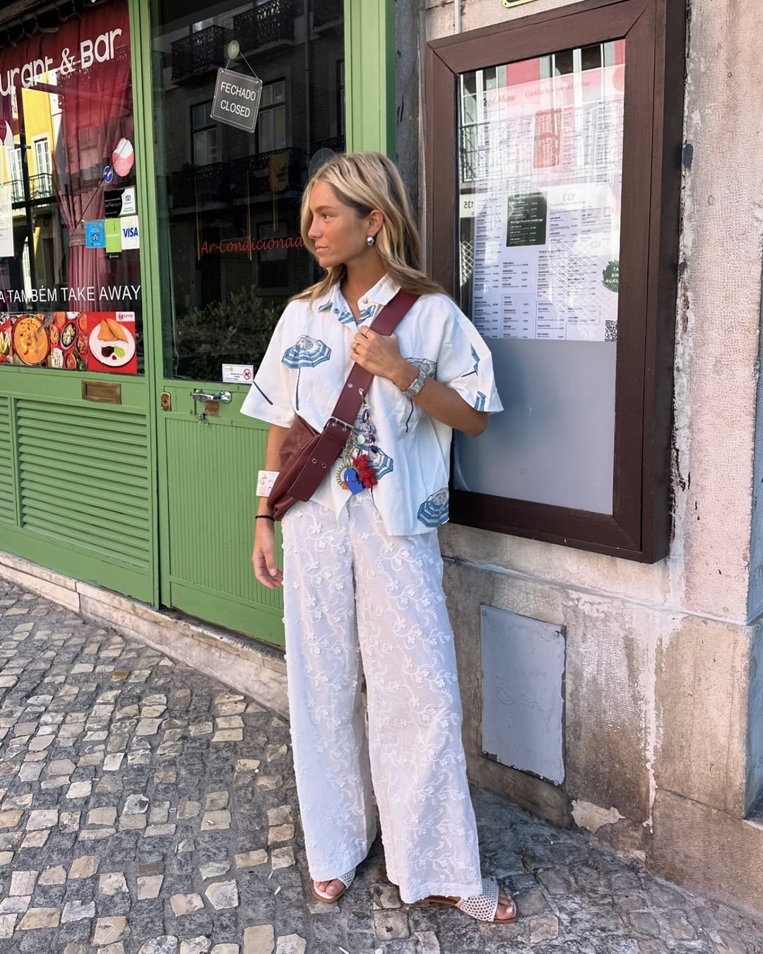 Mulher usa uma camisa oversized estampada e calça larga de renda branca, complementada por sandálias e bolsa transversal marrom com chaveiros.