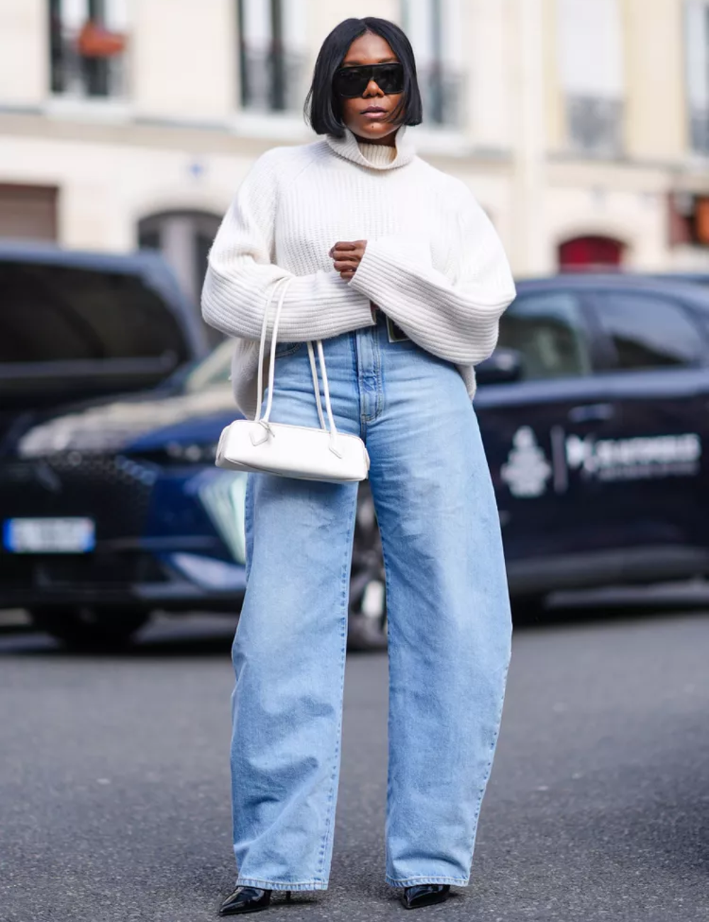 Pessoa vestida com suéter branco oversized, calça jeans wide leg e sapatos de bico fino pretos. Completa o look com bolsa pequena branca e óculos de sol grandes. Combinação moderna de conforto e estilo casual-chique. Tendência urbana outono-inverno 2023.