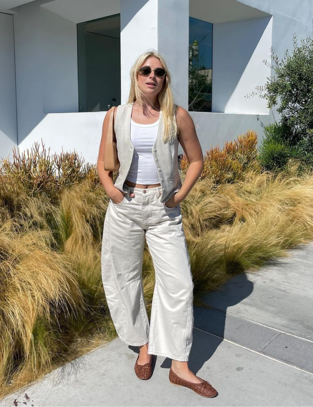 Mulher usando colete e camiseta branca cropped, calça bege larga e sapatilhas marrons. Óculos de sol redondos completam o look casual e confortável, seguindo tendências minimalistas e despojadas da moda urbana.