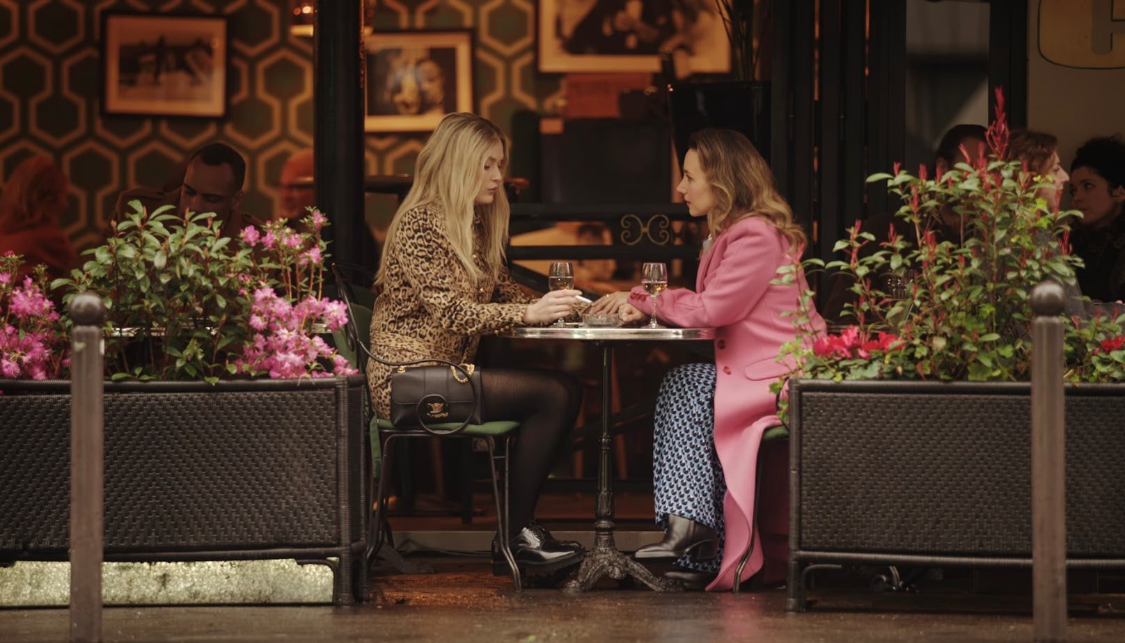 Duas mulheres sentadas em um café, conversando. À esquerda, uma veste um casaco estampado de oncinha, meia-calça preta e bolsa preta.