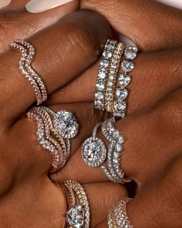 Imagem de close-up de mãos com vários anéis de luxo cravejados de diamantes. As joias apresentam designs modernos e elegantes, incluindo pedras em formatos variados e combinações de metais. Tendência de moda em acessórios sofisticados e exclusivos.