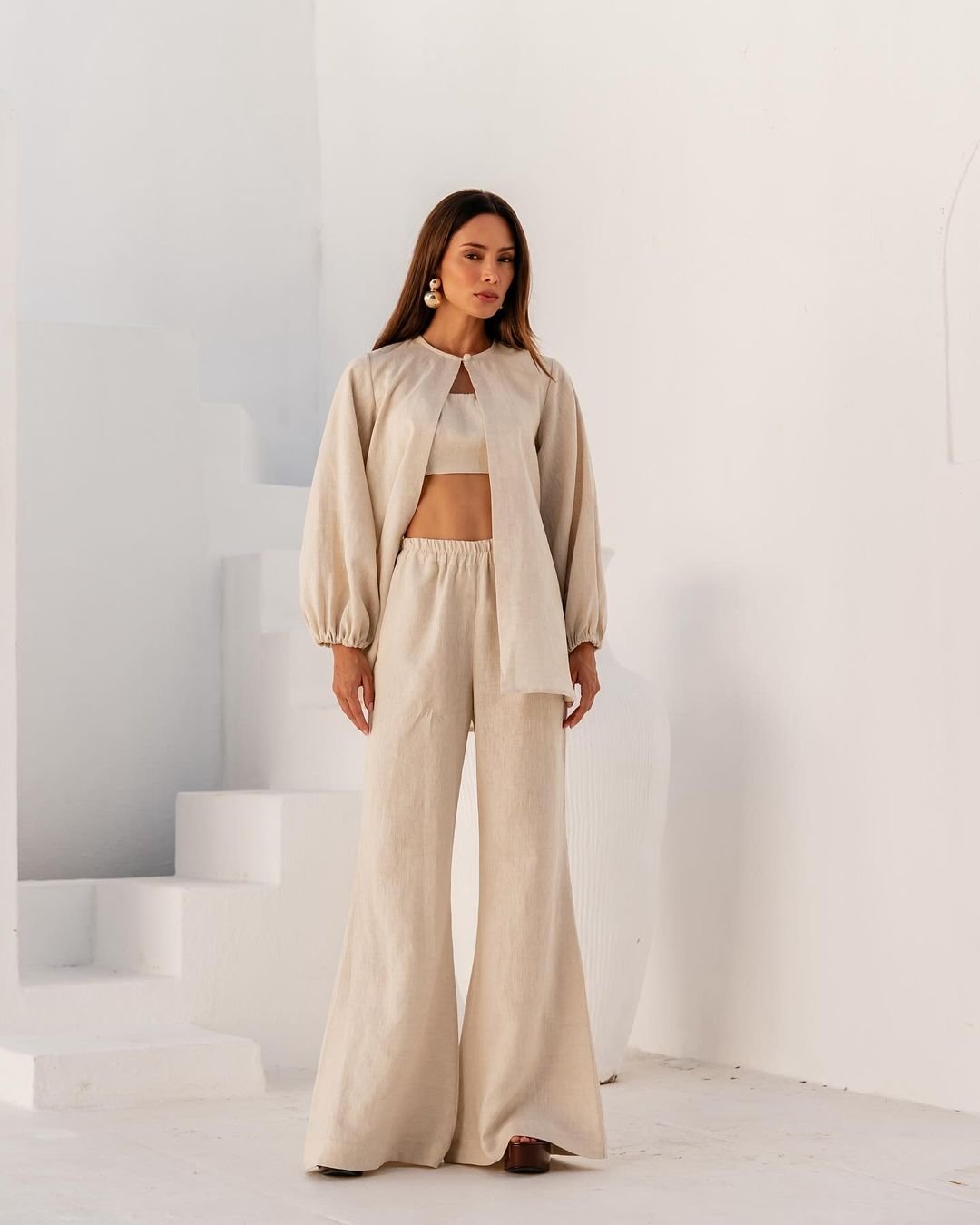 Conjunto feminino em linho bege: top curto, pantalona de cintura alta e casaco com mangas bufantes. Look minimalista e elegante, combinando conforto e sofisticação, perfeito para ocasiões casuais chiques. Tendência de moda para primavera/verão.