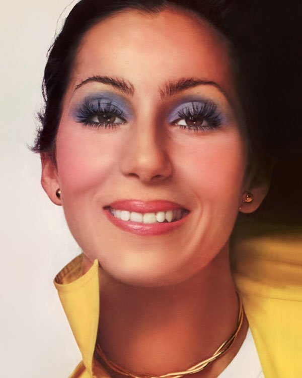 Imagem de mulher com maquiagem marcante: sombra azul vibrante e cílios destacados. Penteado simples com cabelo preso. Usa colar de fios dourados e brincos pequenos. Casaco amarelo, gola levantada. Estilo retrô, anos 70.