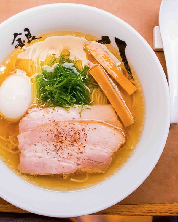 Imagem de um autêntico ramen japonês em uma tigela branca decorada com caracteres japoneses. O prato inclui macarrão, fatias de carne, ovo cozido, vegetais e pedaços de bambu, em um caldo aromático.