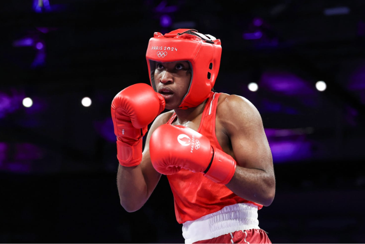 Boxeador nos Jogos Olímpicos Paris 2024 usando uniforme vermelho com luvas e capacete de proteção coordenados. O estilo esportivo é moderno e funcional, garantindo segurança e desempenho. O uniforme também destaca logotipos oficiais, refletindo tendê