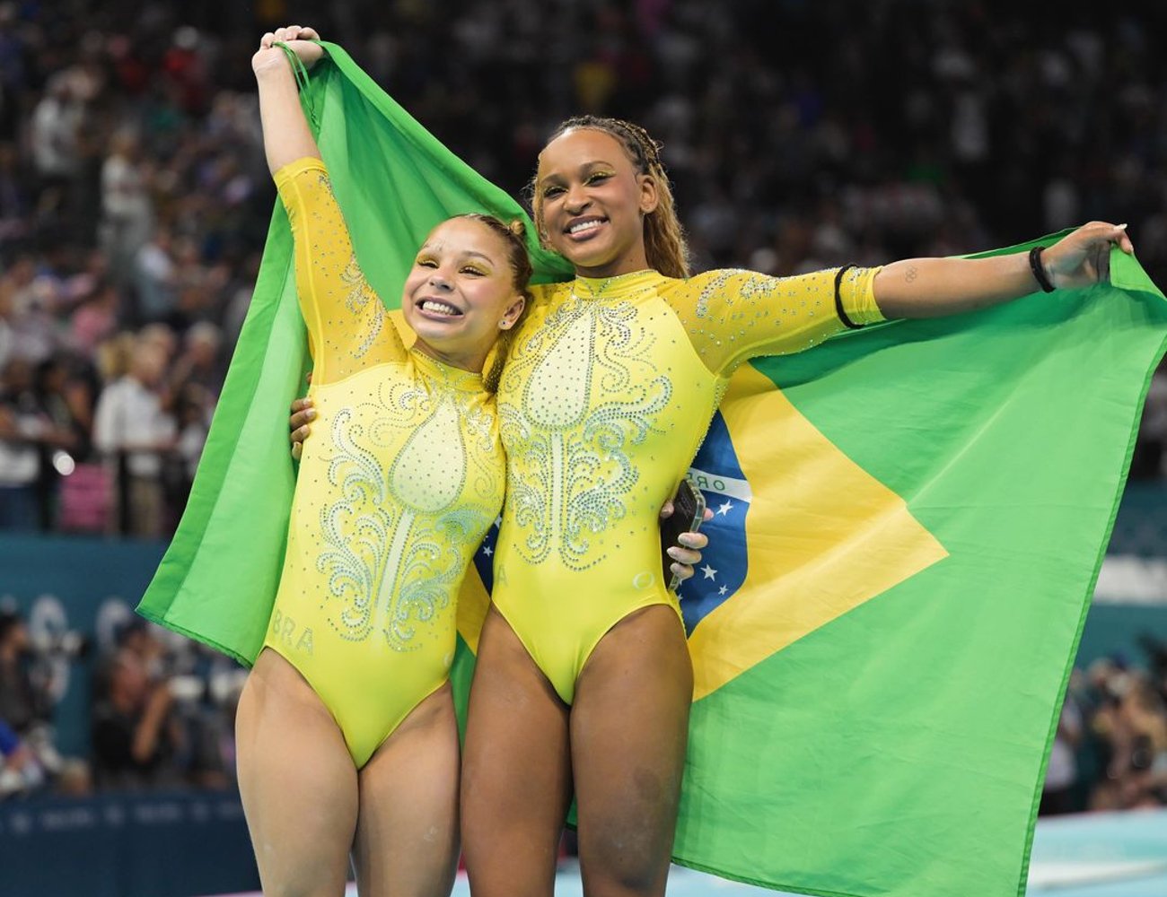 Ginastas brasileiras vestidas com collants amarelos cobertos de pedras decorativas em tons de azul, exibindo com orgulho a bandeira do Brasil. Look combina tendências esportivas e patrióticas com toques de brilho.