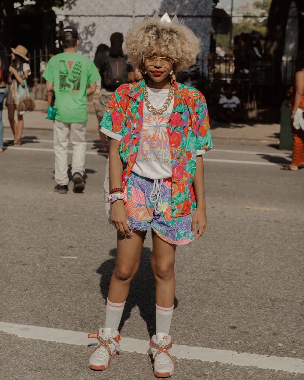 Pessoa com cabelo ondulado e volumoso usando roupas coloridas e estampadas. Ela veste uma camiseta com estampa floral, shorts estampados, camisa desabotoada com cores vibrantes.