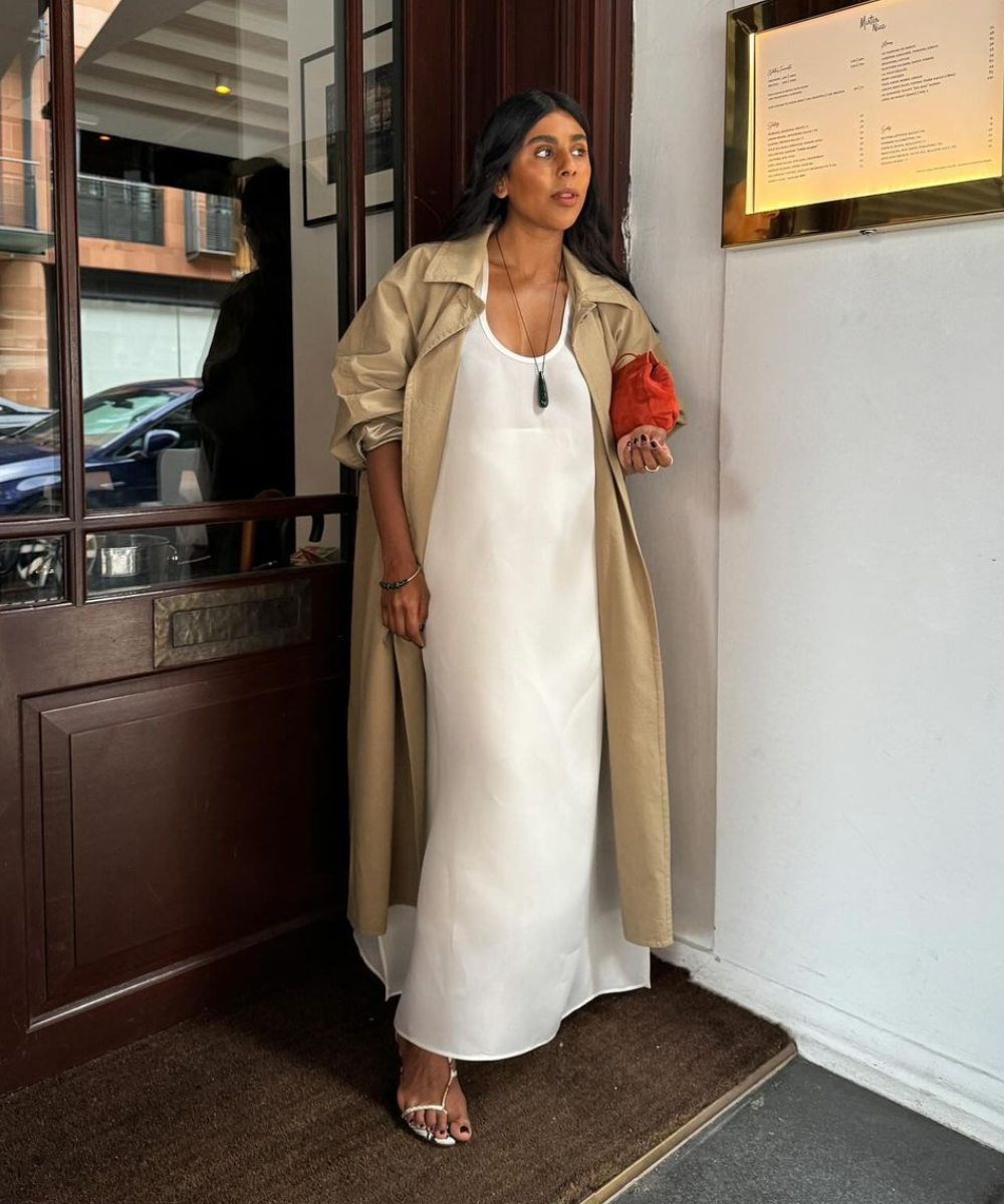 Mulher usando vestido branco longo e esvoaçante, combinado com trench coat bege. Acessórios incluem colar comprido e bolsa clutch laranja. Sandálias minimalistas completam o look elegante. Tendência de moda para 2023: minimalismo e sofisticação casual.