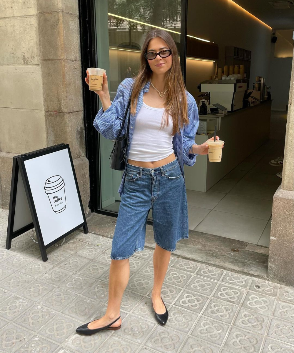 Mulher de cabelo longo e liso usando uma camisa listrada azul sobre uma regata branca e shorts jeans estilo bermuda. Ela complementa o look casual com óculos de sol modernos, uma bolsa preta e sapatilhas pretas, trazendo um estilo urbano e descontraído.