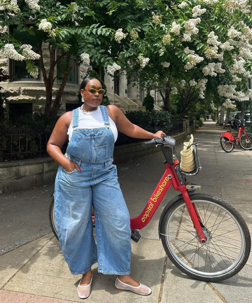 Mulher usando jardineira jeans larga sobre camiseta branca, óculos de sol estilo retrô e sapatilhas transparentes, destacando tendências casuais e confortáveis em um passeio de bicicleta. A moda descontraída é ideal para ambientes urbanos e atividade