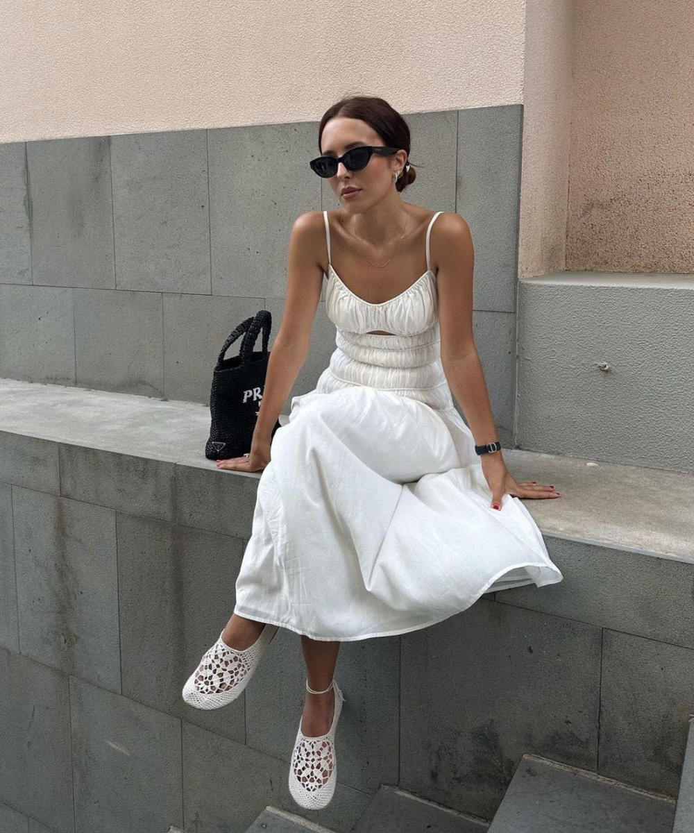 Mulher com vestido branco de alças finas, óculos de sol escuros e sapatos de crochê brancos. Look casual e elegante, ideal para o verão. Roupa complementada com bolsa preta de grife. Tendência minimalista, conforto e estilo.