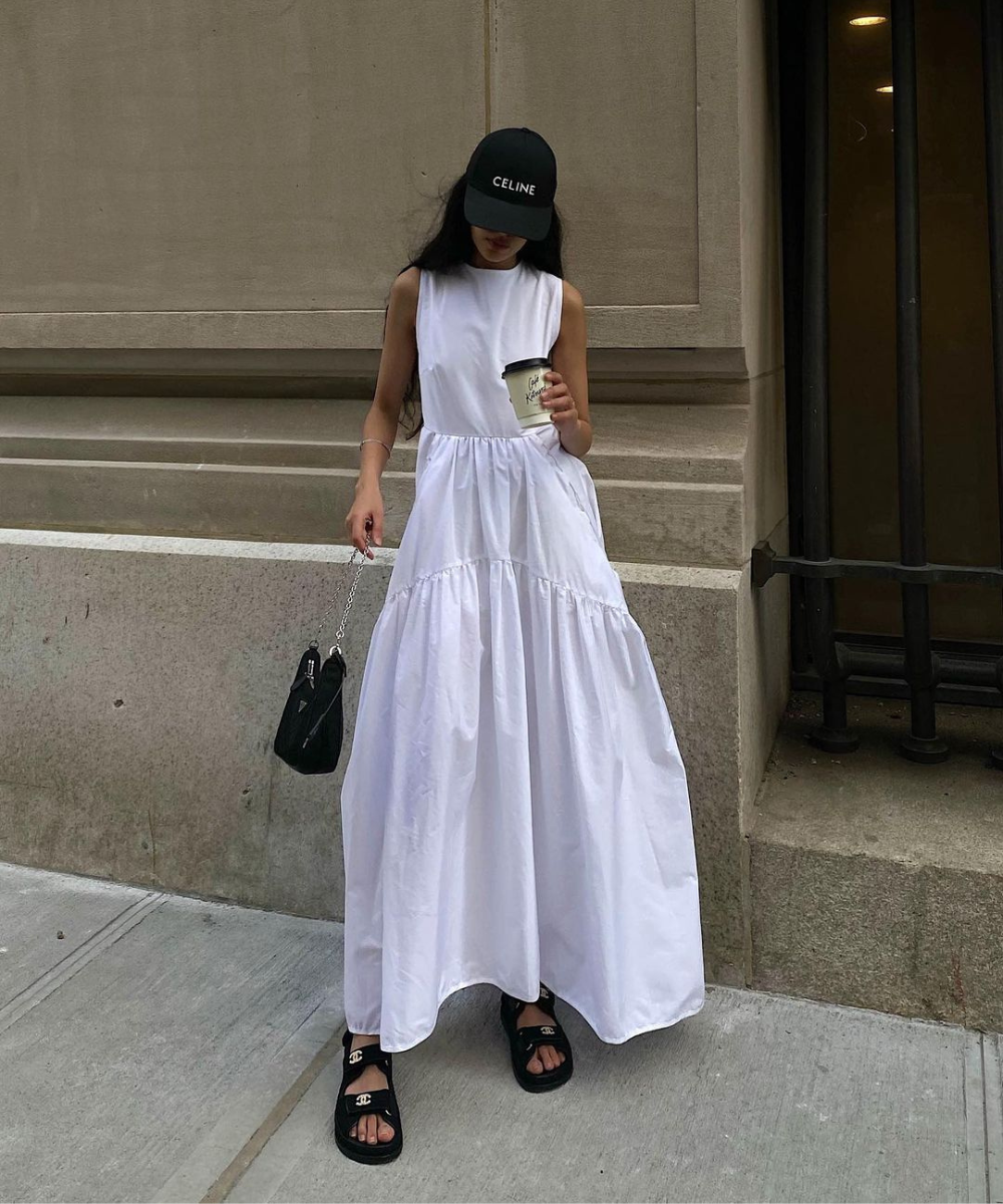 Mulher vestindo vestido longo branco sem mangas, boné preto com logotipo, sandálias pretas e carregando bolsa preta. Estilo casual e effortless chic, combinando conforto com elegância minimalista.