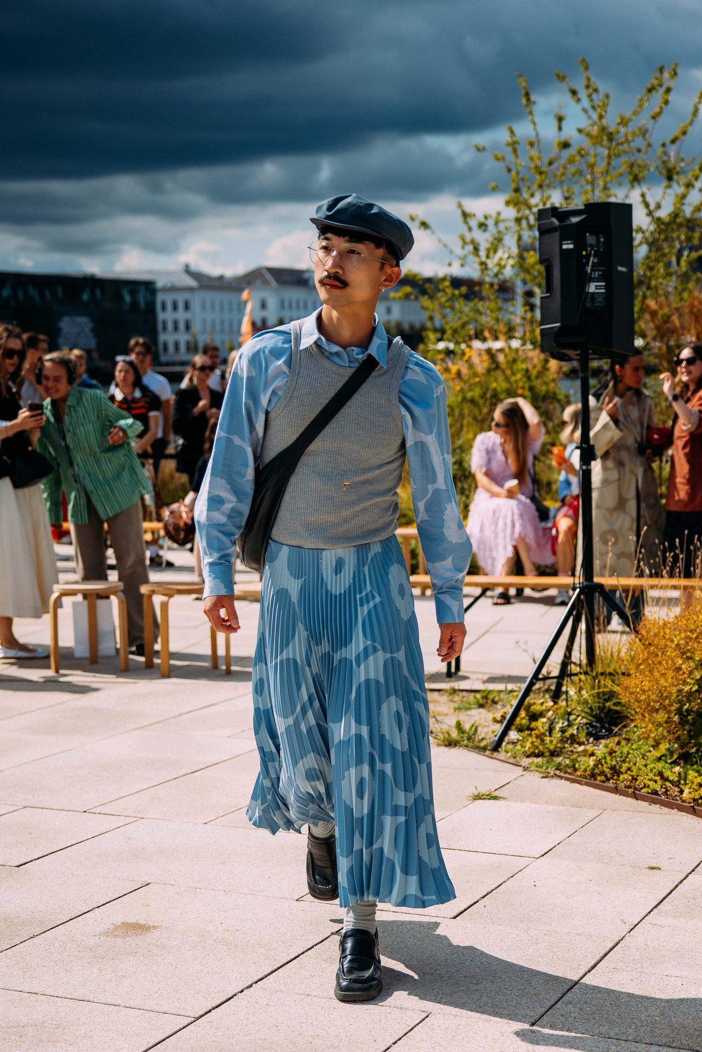 Homem com look fashionista, combinando boina azul, camisa estampada, colete cinza e saia plissada azul com padrão geométrico. Tendência de moda genderless, mixando peças clássicas e modernas, destacando estilo pessoal e ousadia no uso de acessórios.