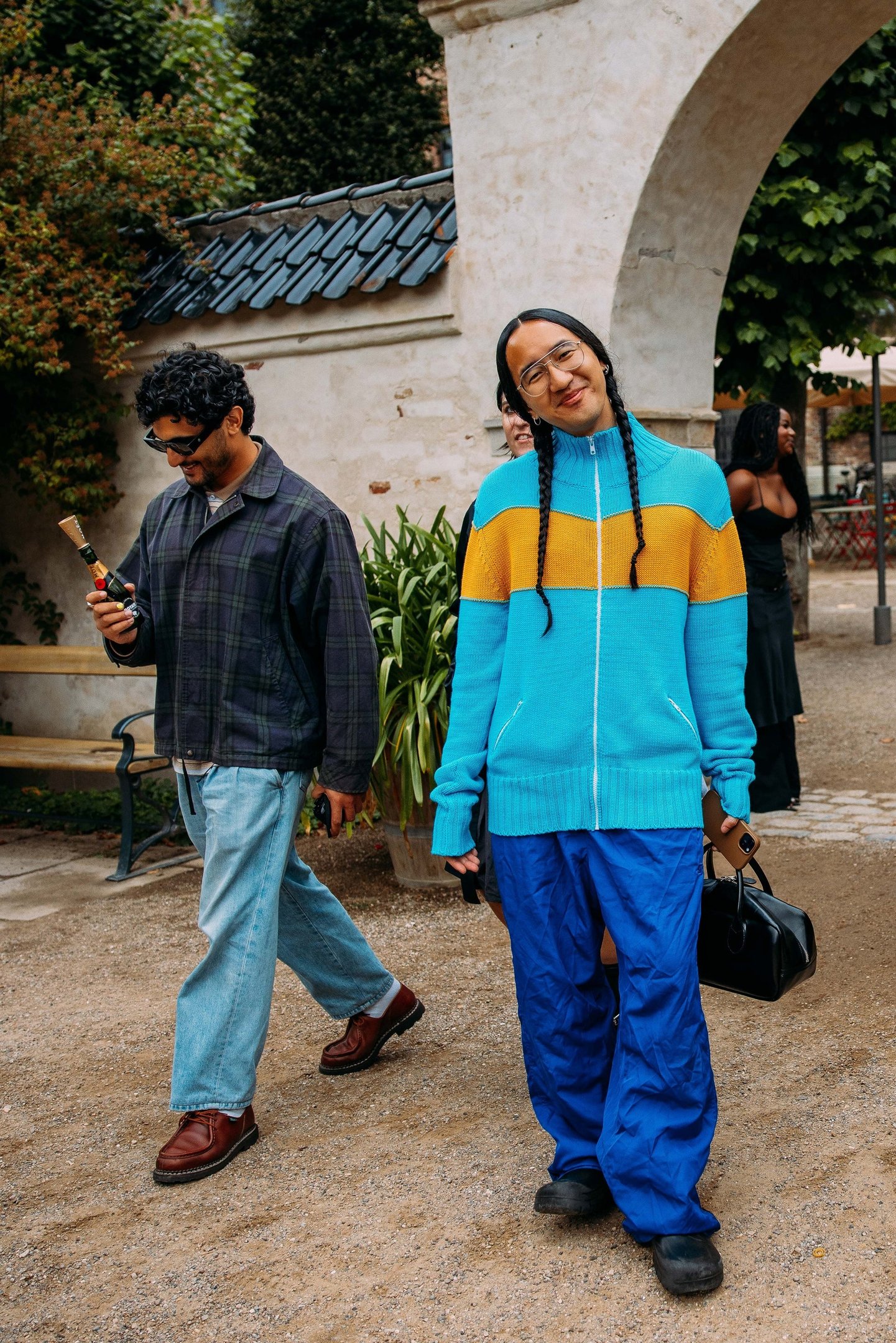 Pessoas com estilo casual e despojado. À esquerda, homem com jaqueta xadrez, jeans largos e sapatos marrons. À direita, pessoa com camiseta azul e laranja, calça esportiva azul e óculos grandes. Ambos seguem tendências de conforto e mistura de cores vibrantes.