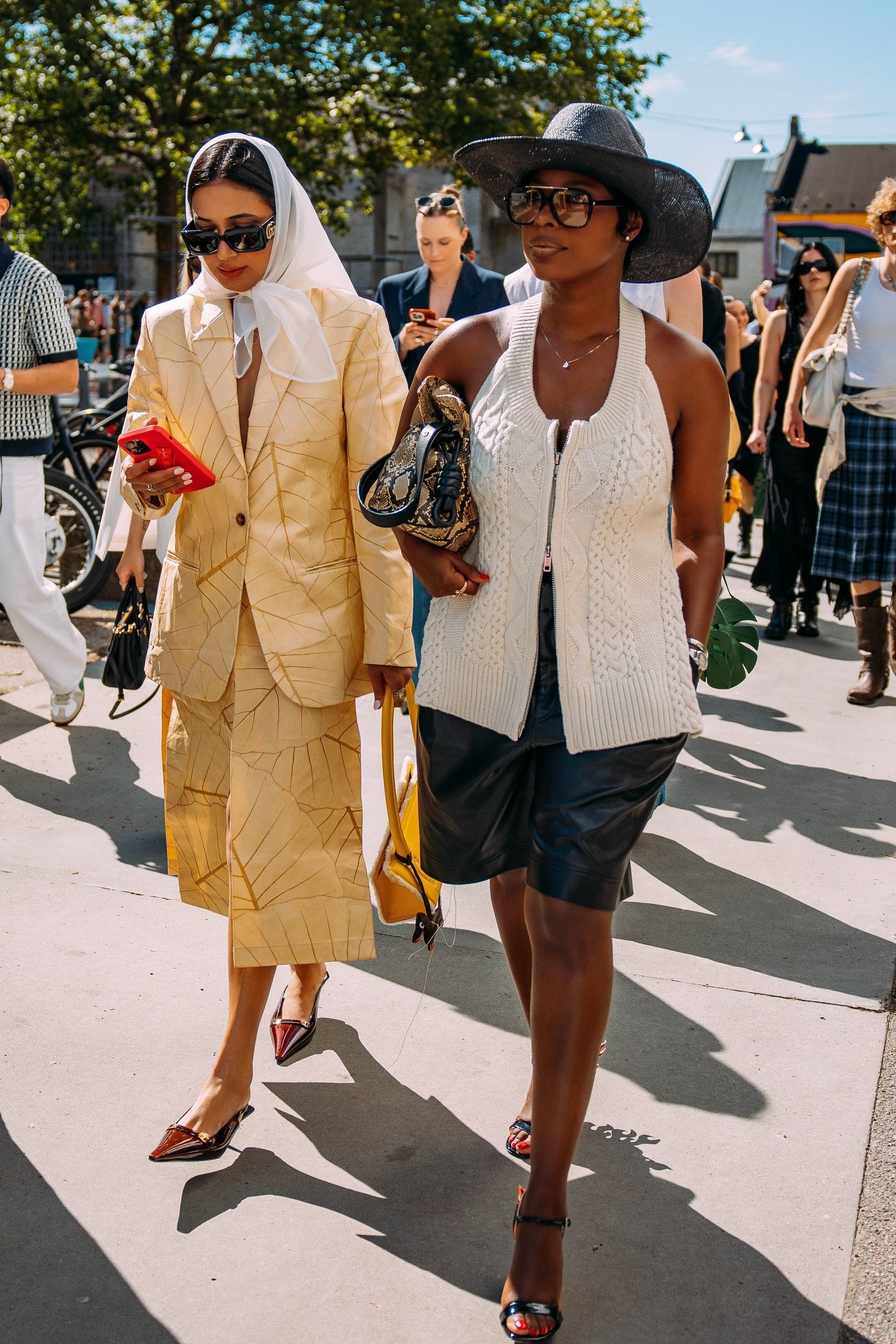 Duas mulheres em destaque: uma usando conjunto bege com casaco e saia midi, com lenço branco na cabeça e bolsa amarela. Outra vestindo colete de tricô branco, bermuda de couro preto, chapéu preto e óculos de sol grandes.