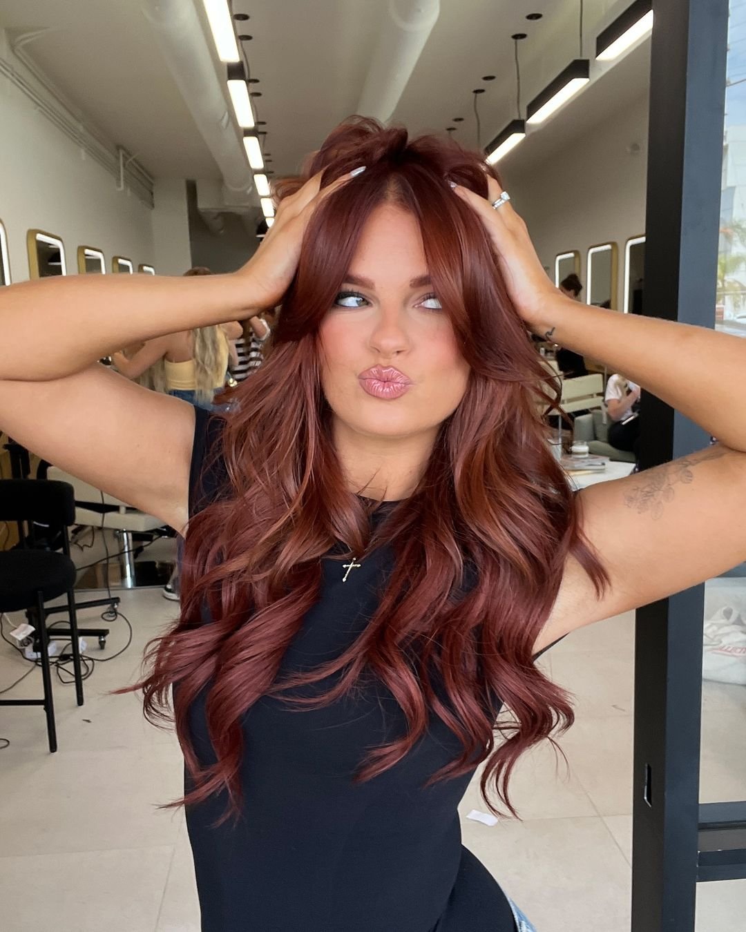 Mulher com cabelo ruivo ondulado, usando vestido preto ajustado, com corrente e crucifixo prateado. Tendências de moda demonstradas: cabelo ruivo vibrante, vestido preto básico e acessórios minimalistas. cores de cabelo tendência