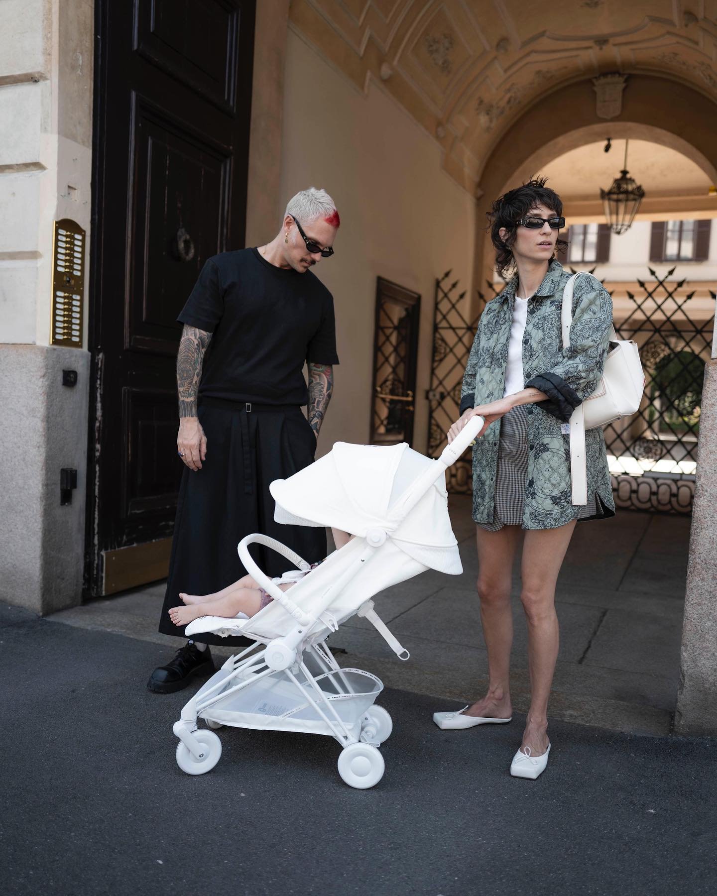 Casal elegante com bebê em carrinho branco. Homem com cabelos loiros curtos e tingidos de vermelho, vestido com camiseta preta e saia longa preta, combinado com óculos escuros.