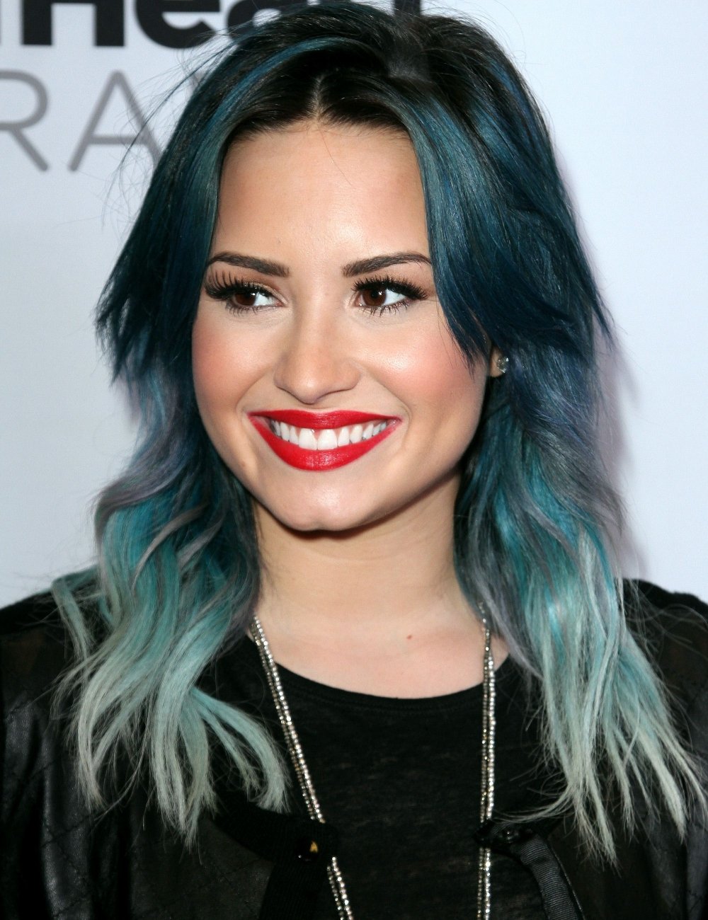 Demi Lovato com cabelo ombré em tons de azul, maquiagem destacada com batom vermelho vibrante e expressão sorridente. Usa um colar prateado e uma roupa preta com textura de couro, refletindo tendências modernas e ousadas de moda.