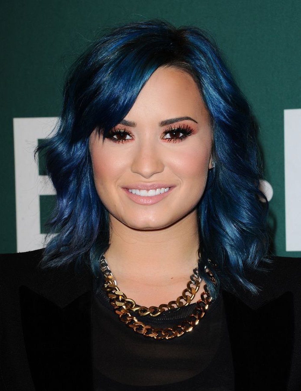 Demi Lovato com cabelo azul vibrante em corte médio e ondulado, maquiagem com olhos esfumados e lábios naturais. Veste blusa preta com sobreposição em tecido transparente e colar de correntes douradas sobre blazer preto.