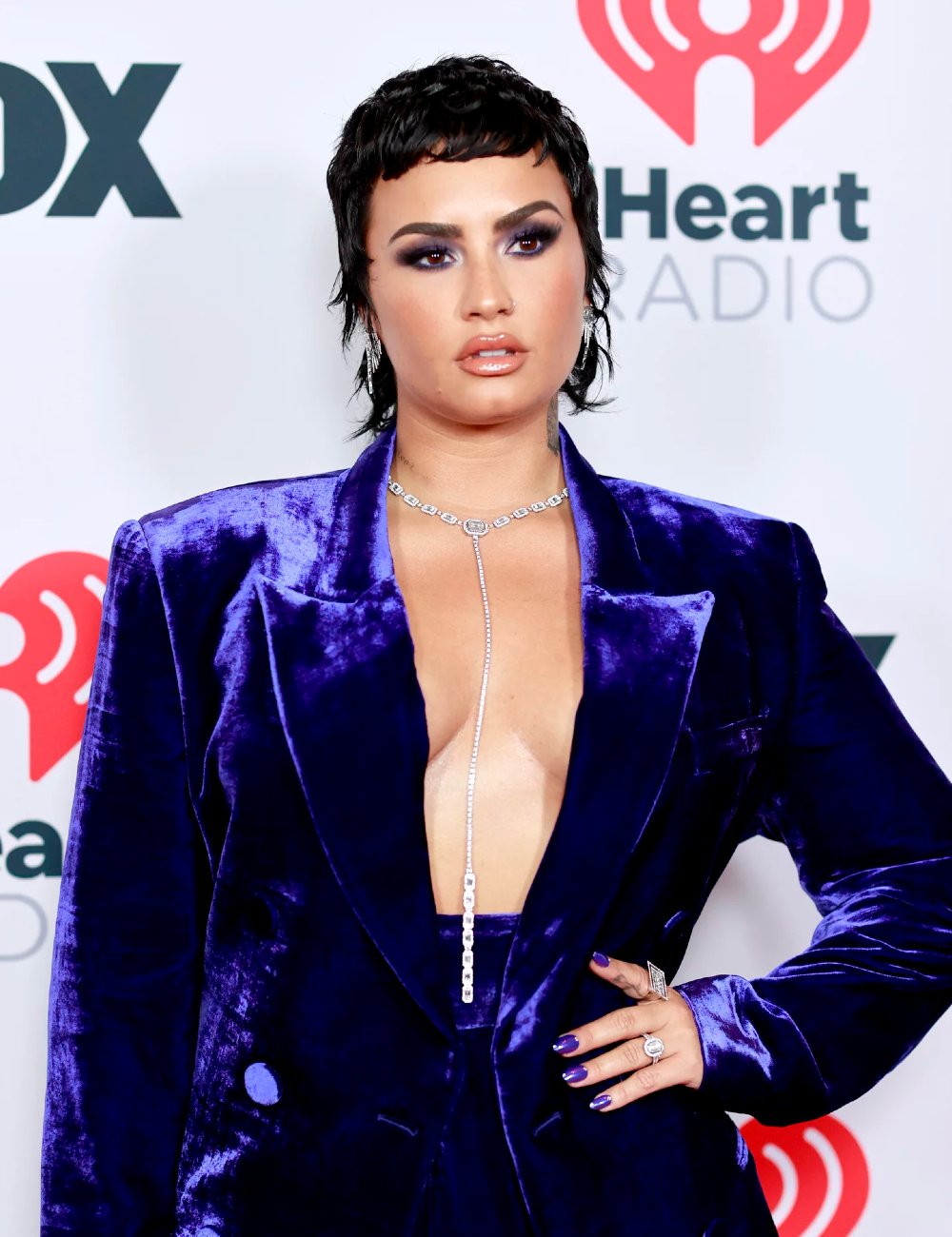 Demi Lovato com cabelo curto e estilizado, maquiagem dramática com sombra escura nos olhos. Unhas combinando com o traje, enfatizando um visual ousado e sofisticado.