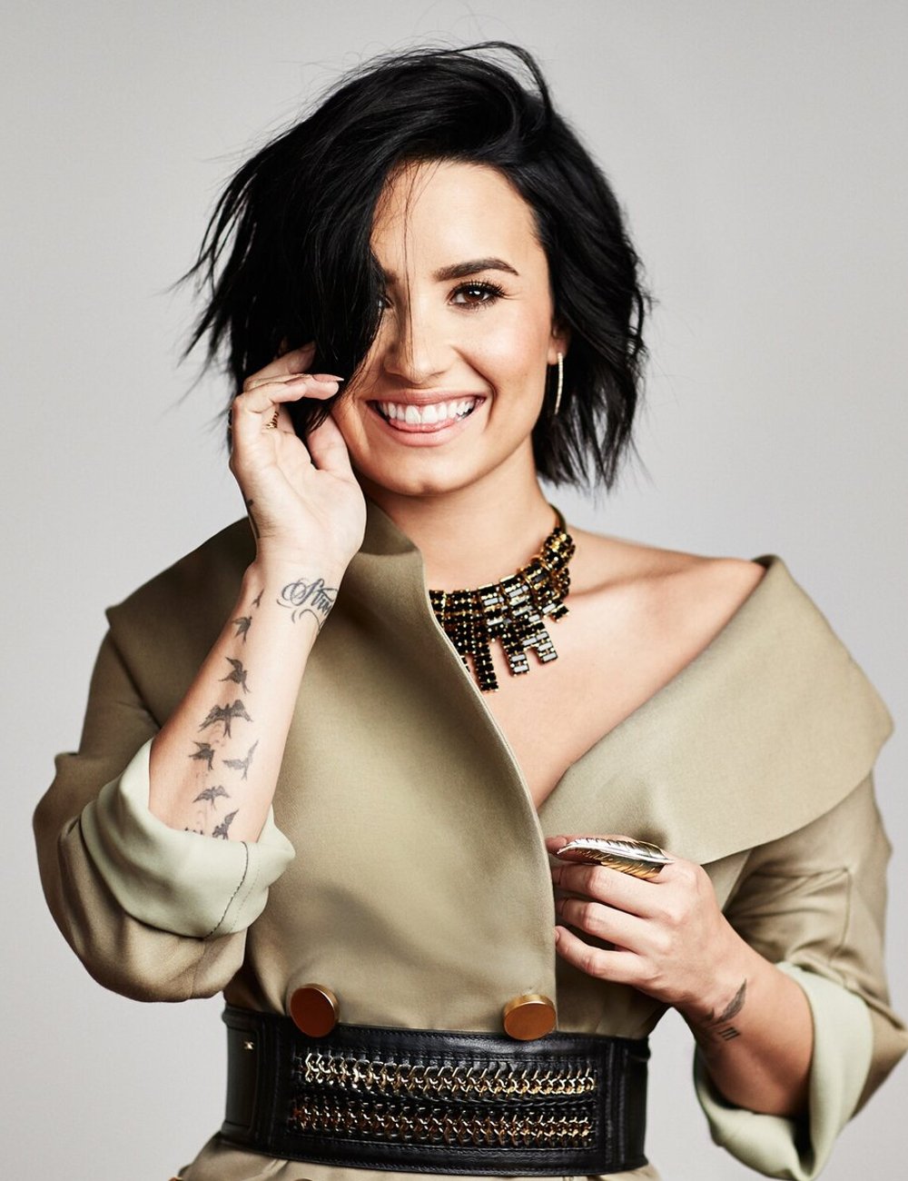 Demi Lovato com cabelo preto curto e moderno, usando um casaco bege oversized e acessorizado com um cinto largo de couro preto e detalhes dourados. No colo, um colar statement de miçangas pretas.