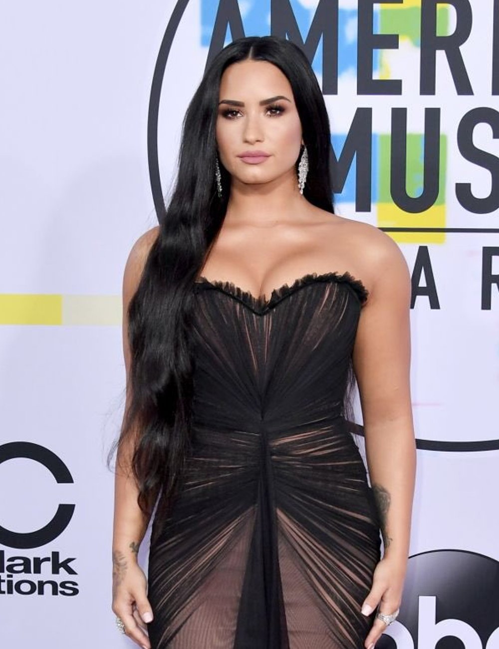 Demi Lovato usando um vestido longo preto com detalhes estruturados de tule e transparências. O look é sofisticado, destacando o busto com um decote tomara que caia. A produção é complementada por cabelo solto e liso e brincos longos prateados.