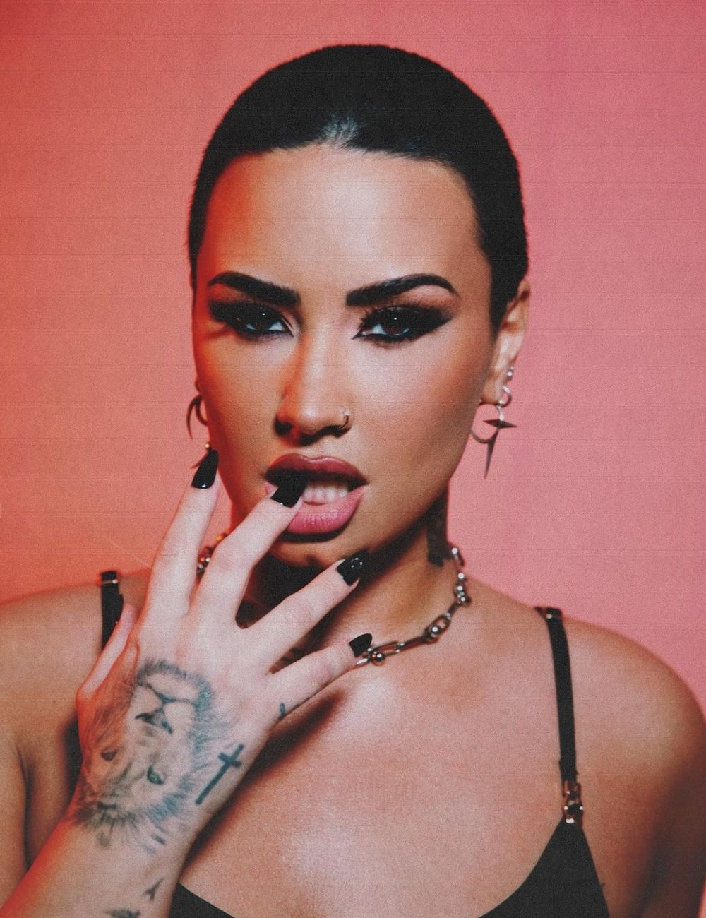 Demi Lovato com cabelo curto e maquiagem marcante, usando brincos longos, colar estilo gótico e alças finas no top. Tatuagem de leão na mão esquerda e unhas compridas escuras. Tendências de moda alternativa, gótica e punk.