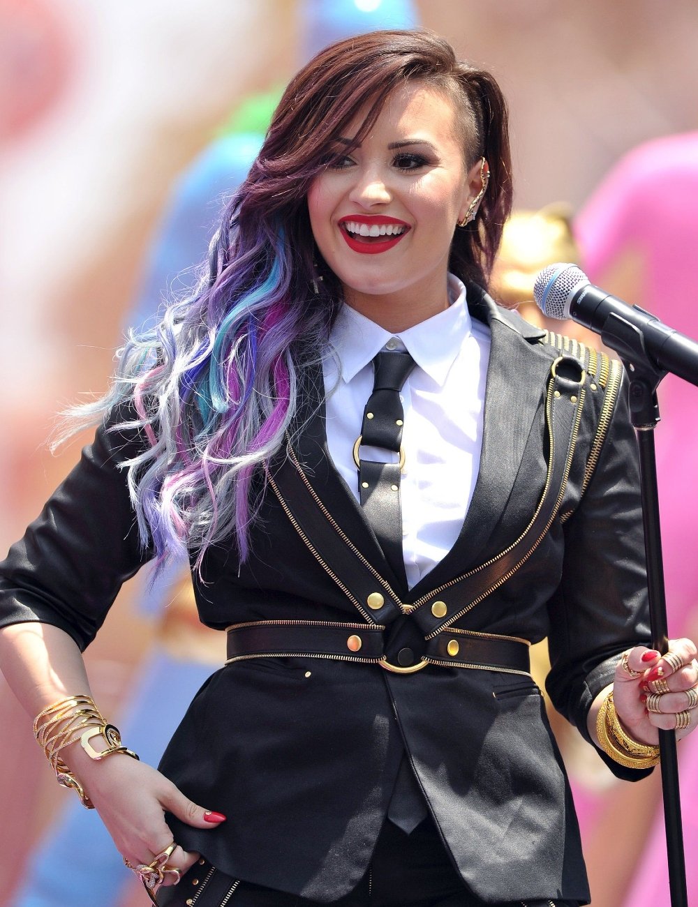 Demi Lovato vestindo blazer preto com detalhes dourados, camisa branca e gravata preta elegante. Cabelo longo e colorido em tons de roxo e azul. Acessórios chamativos, como pulseiras douradas e lipstick vermelho.