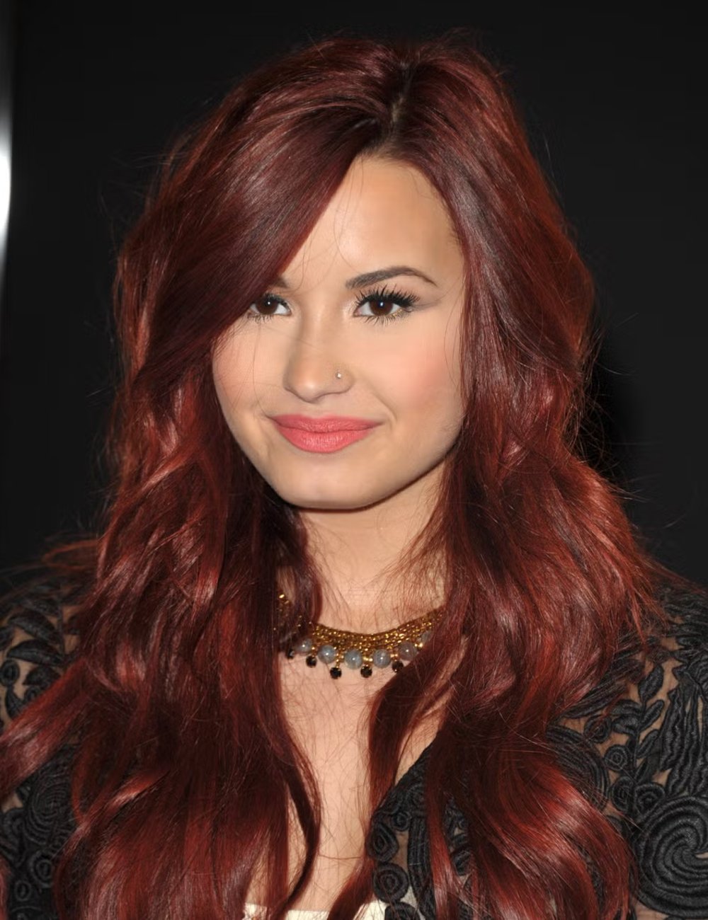 Demi Lovato com cabelo vermelho ondulado em tom vibrante, combinando com maquiagem de destaque nos olhos e batom suave. Colar de pedras harmoniza com o vestido preto rendado, ressaltando uma expressão de moda elegante e moderna.