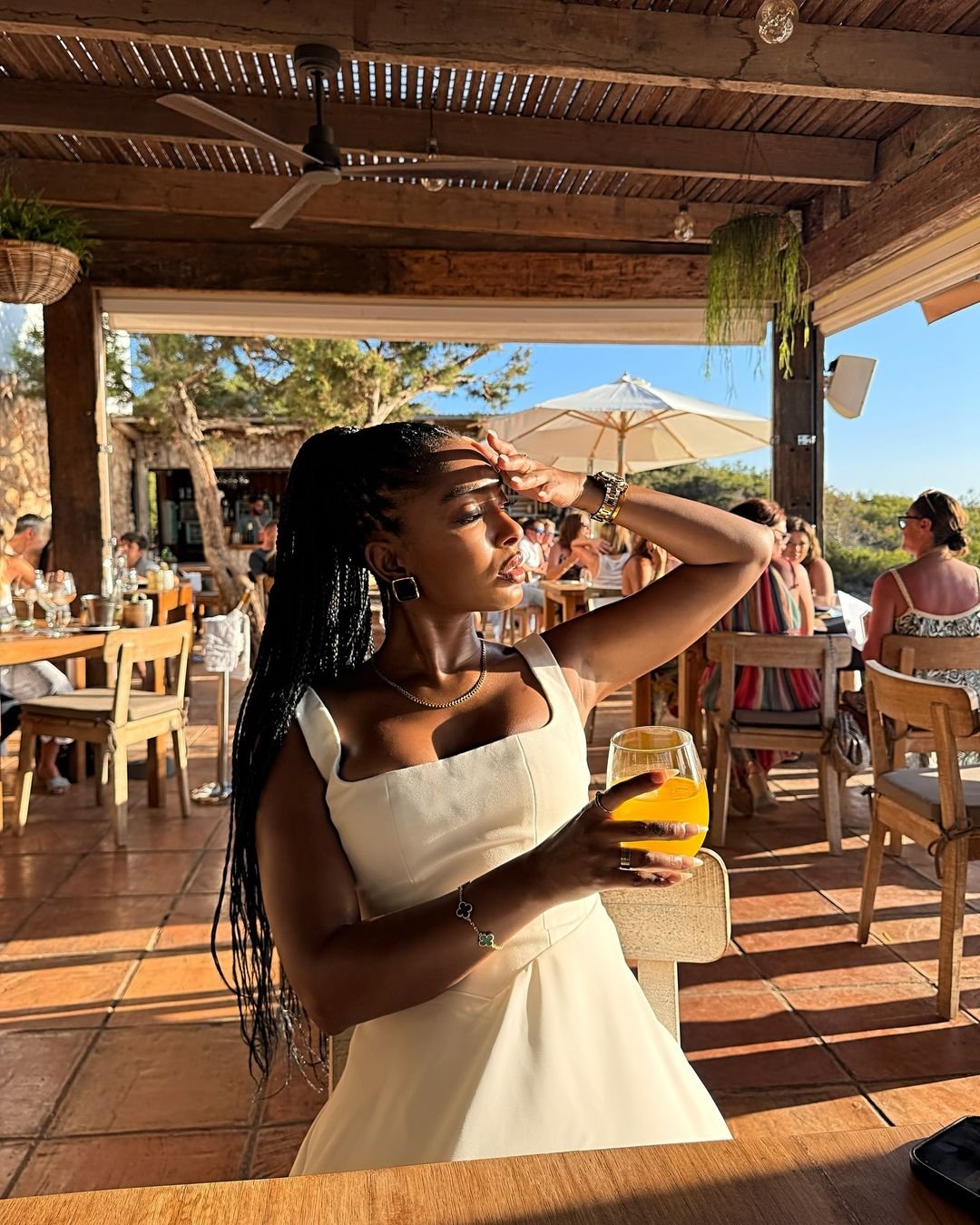 Mulher elegante em um restaurante ensolarado, vestindo um vestido branco minimalista e acessórios discretos. Tendências de verão com toque sofisticado, mostrando tranças longas e impecáveis, desodorante com niacinamida