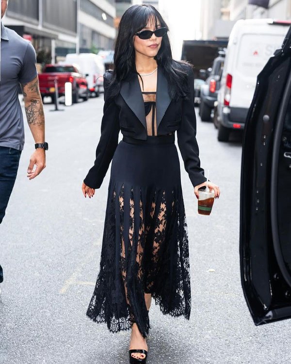 Mulher com look elegante: Vestido preto com saia de renda transparente, blazer curto com decote profundo, óculos escuros e sandálias de salto. Tendência de moda urbana chique, misturando sofisticação e sensualidade com toque moderno.