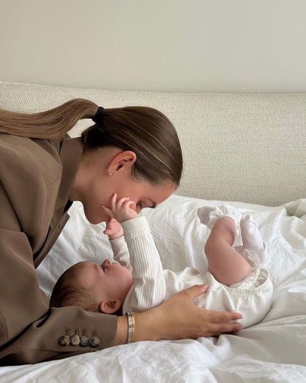 Mulher usando blazer marrom e rabo de cavalo brinca com bebê vestido de branco. Estilo casual chic com maquiagem minimalista e acessórios discretos. Tendência de moda confortável e atemporal para mães modernas.