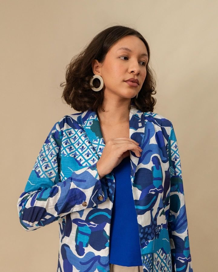 Mulher vestindo um blazer estampado em tons de azul e branco, combinando com uma blusa azul. Acessório marcante: brincos de argola grandes e circulares. Look contemporâneo e vibrante expressando tendências modernas e criativas de moda casual chic.