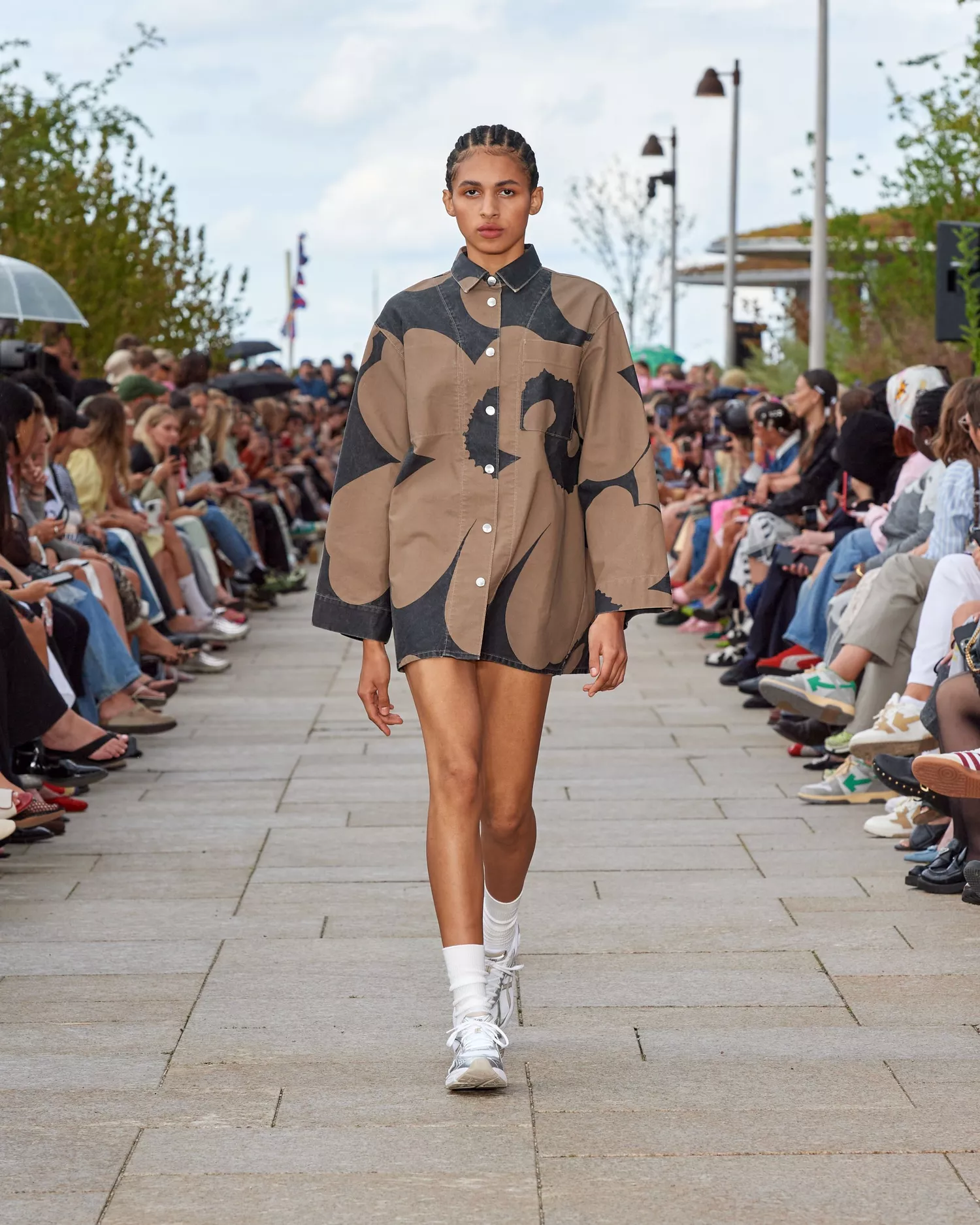 Desfile de moda com foco em tendências contemporâneas. Modelo veste uma camisa oversized com estampa floral em tons neutros, combinada com shorts escuros.