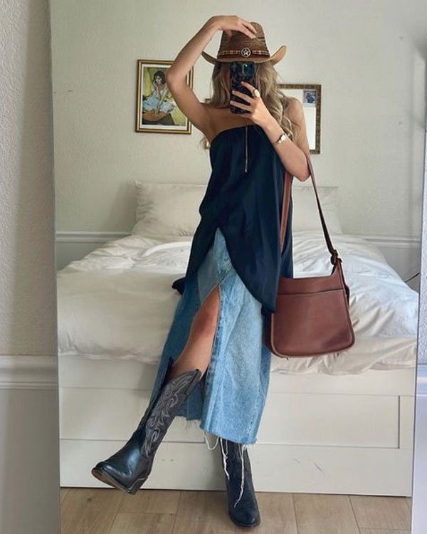 Mulher vestindo chapéu estilo cowboy, blusa preta tomara que caia, saia longa jeans com fenda frontal, botas de cowboy pretas e bolsa marrom transversal. Look combina elementos do estilo boho e western.