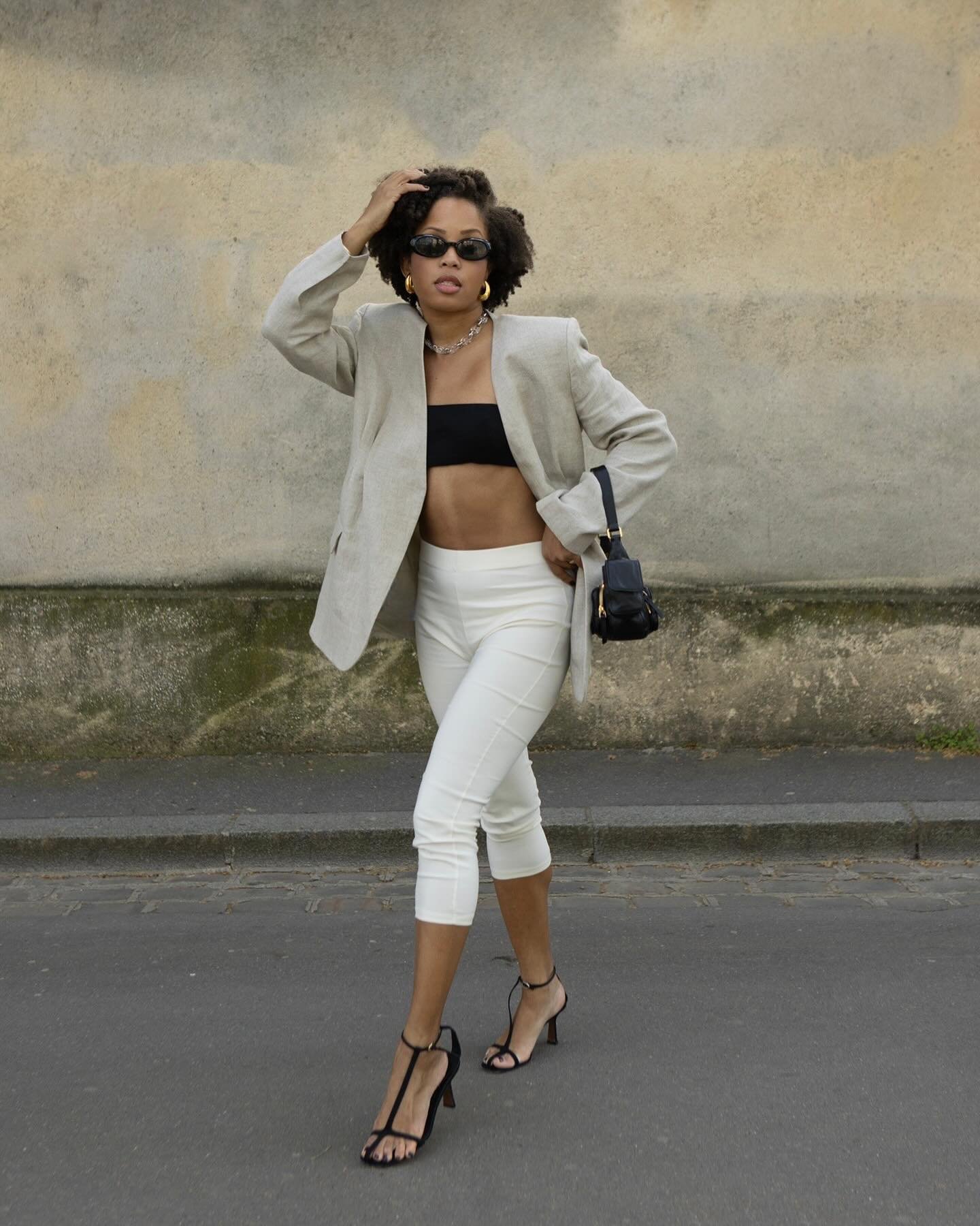 Mulher com blazer bege oversized, top preto, calça branca capri justa e salto alto preto, carregando uma bolsa pequena. Óculos de sol e colar chunky completam o look moderno e sofisticado, tendência de moda atual urbana e chic.