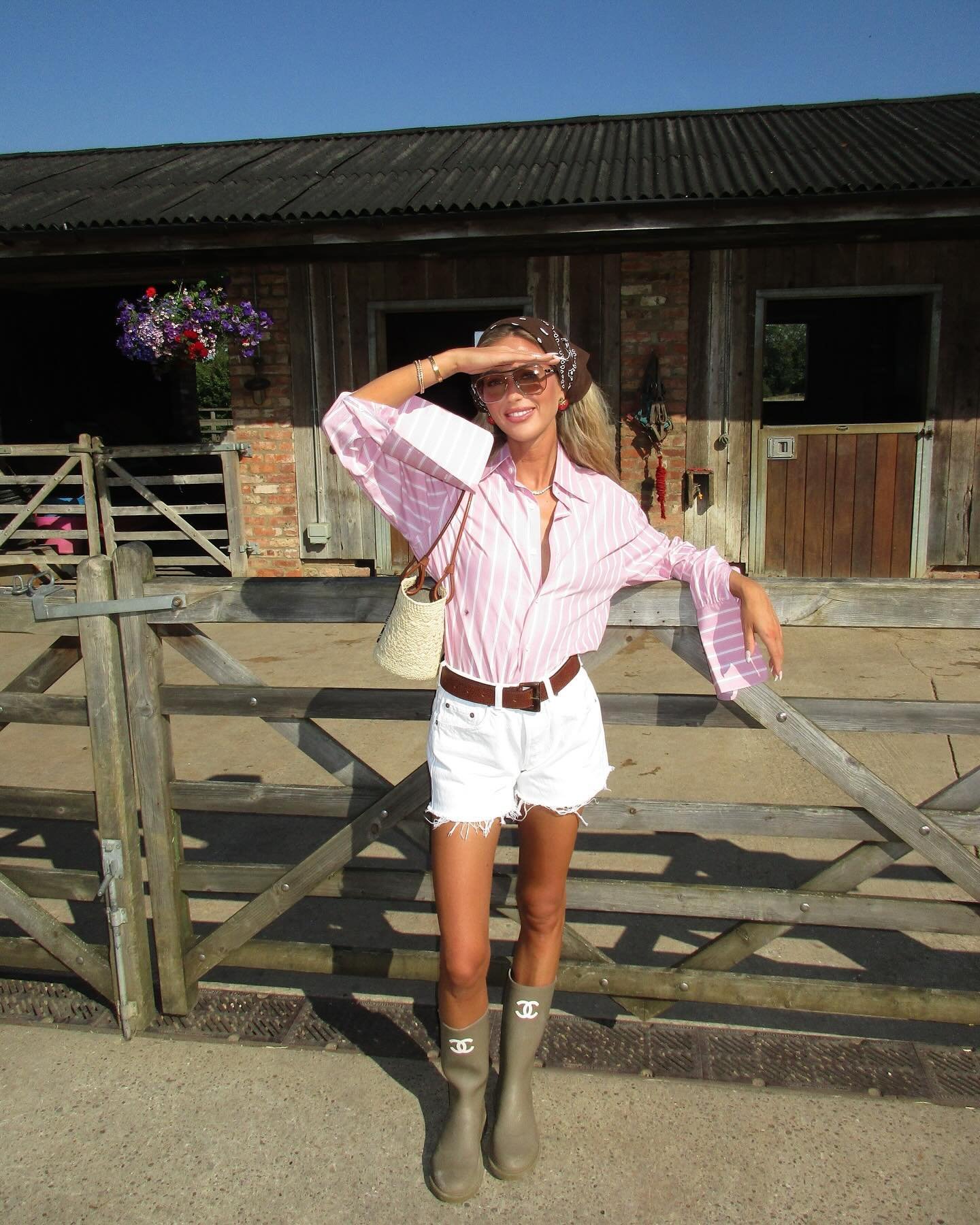 Pessoa vestindo camisa listrada rosa e branca com short branco desfiado. Completa o visual com botas de borracha e cinto marrom, além de uma bolsa de palha e lenço marron de estampa floral.